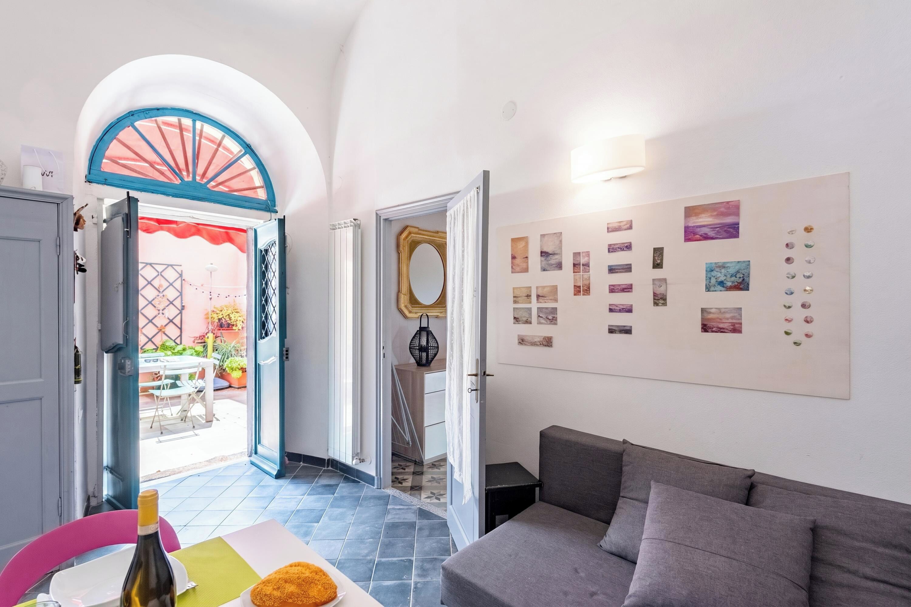 A room with a sofa, a table with a bottle and a plate, a wall with framed pictures, and an open door leading outside.