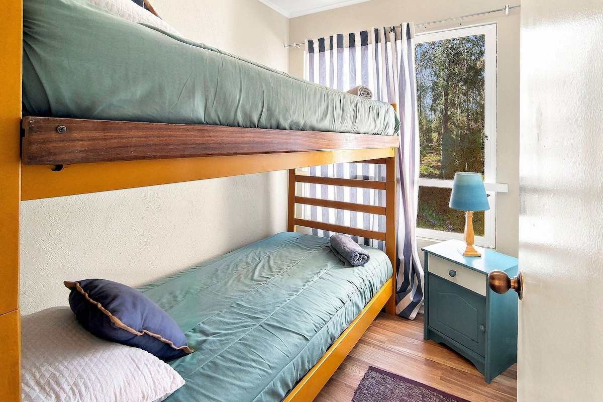 Un lit superposé avec une structure en bois, un lit simple avec un matelas vert, une petite table de chevet avec une lampe et une fenêtre avec des rideaux.