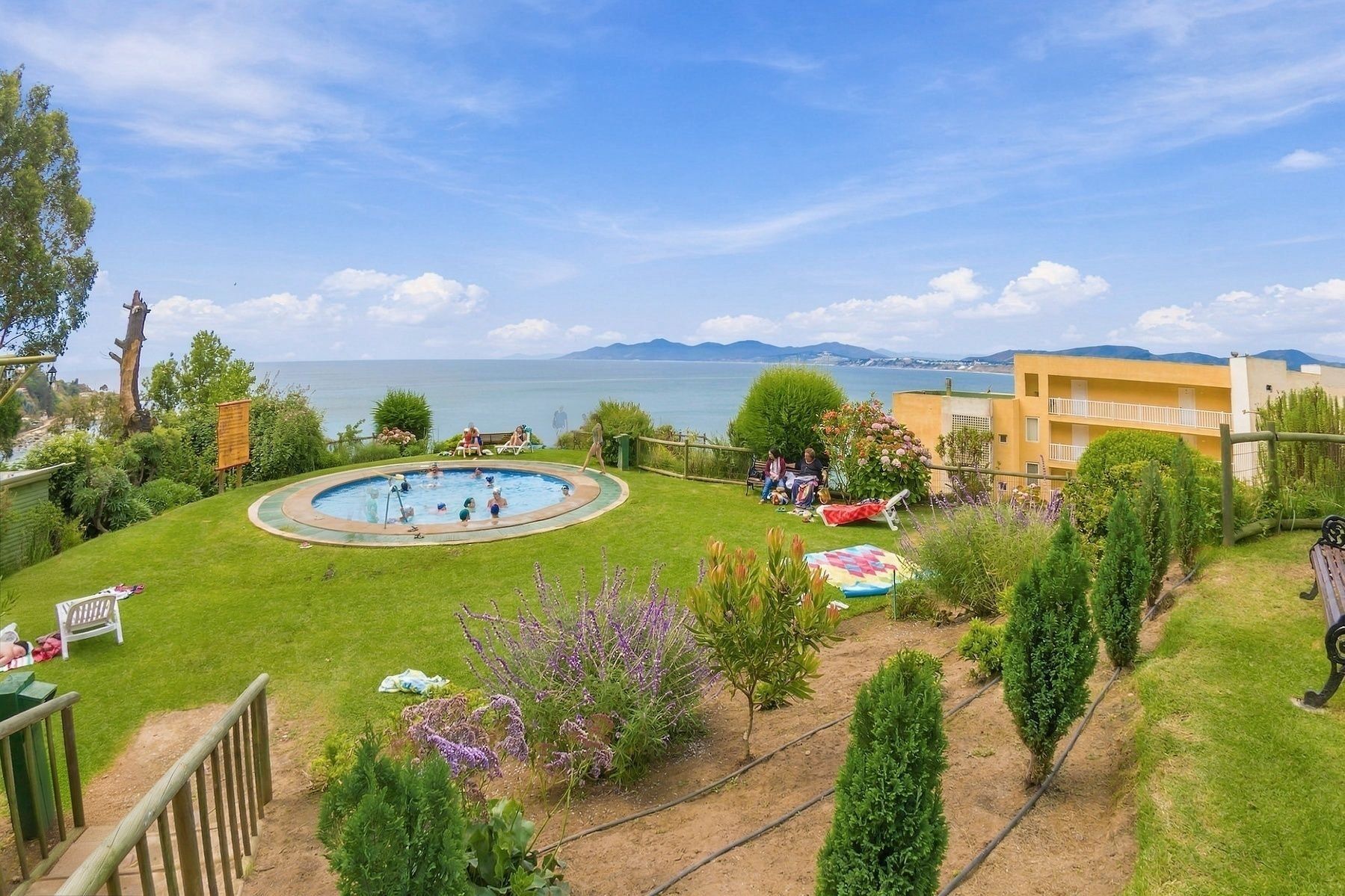 A pool area with a view of the sea, surrounded by greenery and a building.