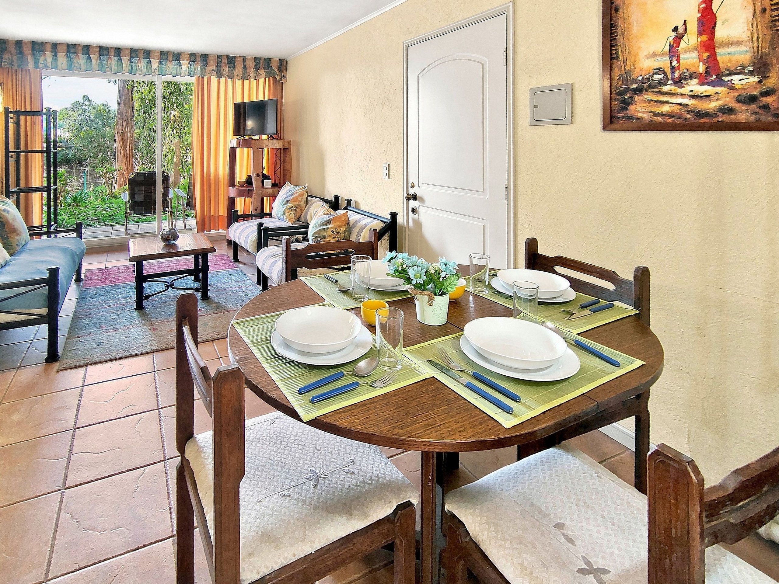Zona pranzo con tavolo apparecchiato per quattro persone, un divano e una televisione sullo sfondo.