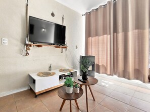 A modern living room with a flat-screen TV mounted on the wall, a white entertainment unit, two small round tables with potted plants, and a large window with curtains. - Oceana Suites Bahía Pelicanos (Puchuncavi)
