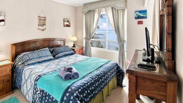 Quarto com cama, mesa de cabeceira, escrivaninha, janela com cortinas e vista para o mar.