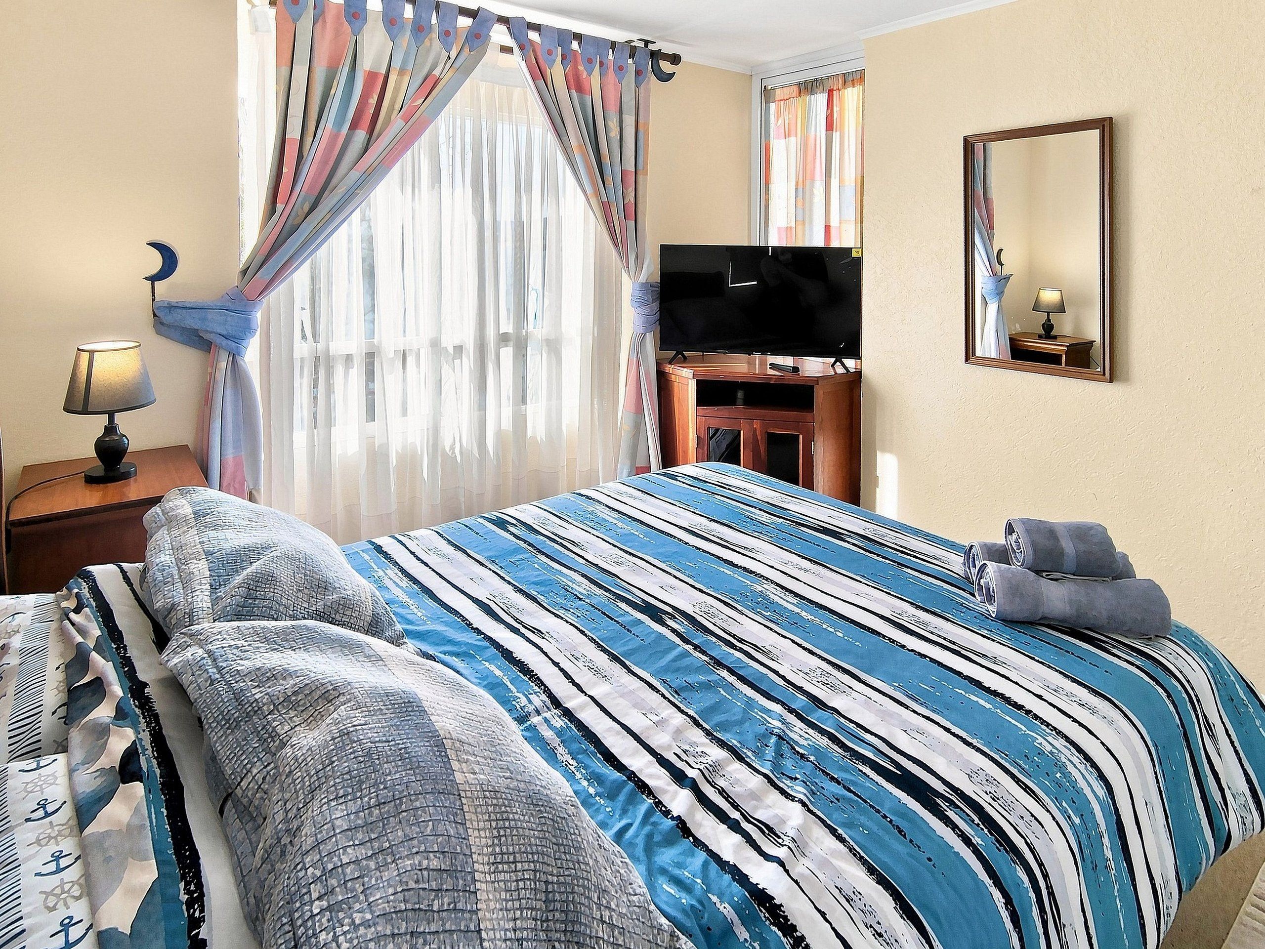 A hotel room with a bed, a television, a mirror, and a nightstand with a lamp.