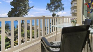 A balcony with a glass table and chairs, overlooking a coastal area with trees and a building in the background.
