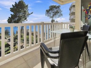 Un balcon avec une table et des chaises en verre, donnant sur une zone côtière bordée d’arbres, avec un bâtiment visible au loin.
