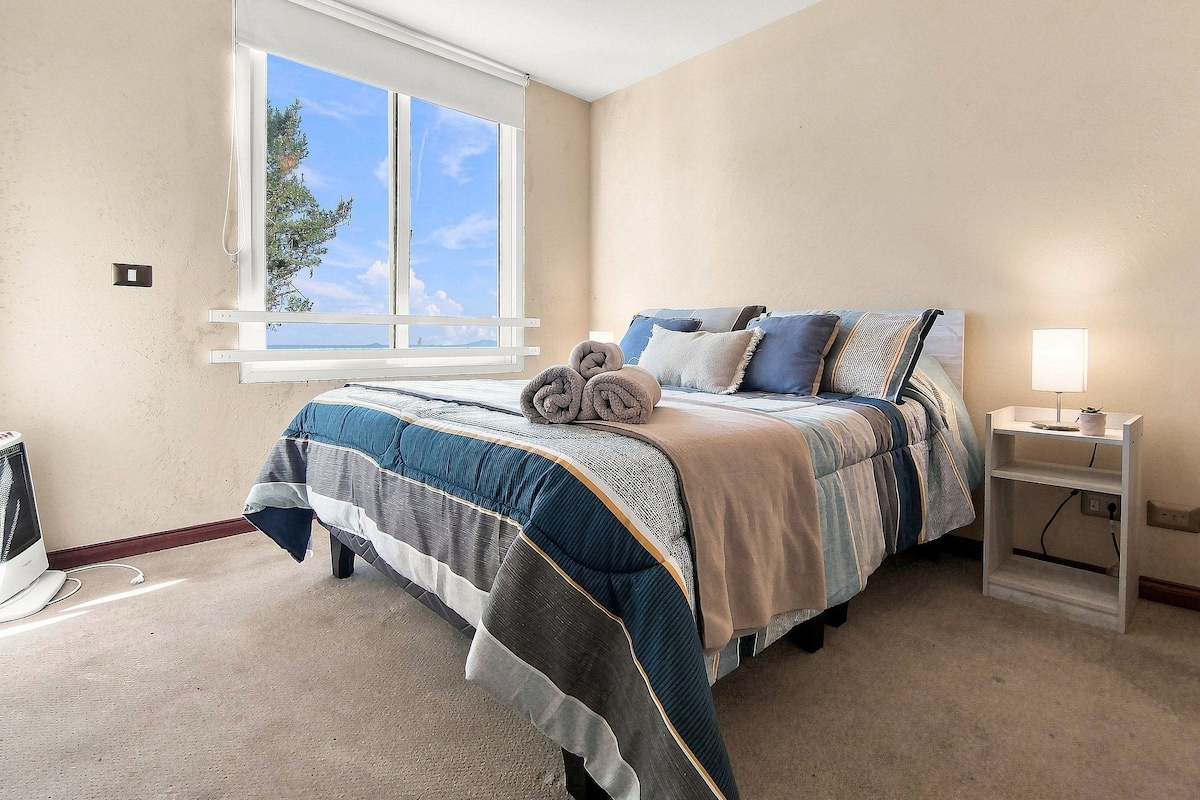 Une chambre avec une grande fenêtre, un lit recouvert d’une courtepointe à rayures, une lampe de chevet et une petite table sur laquelle est posée une plante.