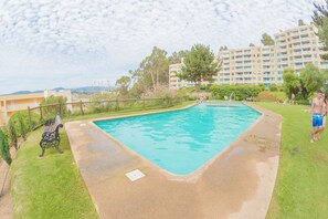 A swimming pool with a grassy area, a bench, and a building in the background. - Oceana Suites Bahía Pelicanos (Puchuncavi)