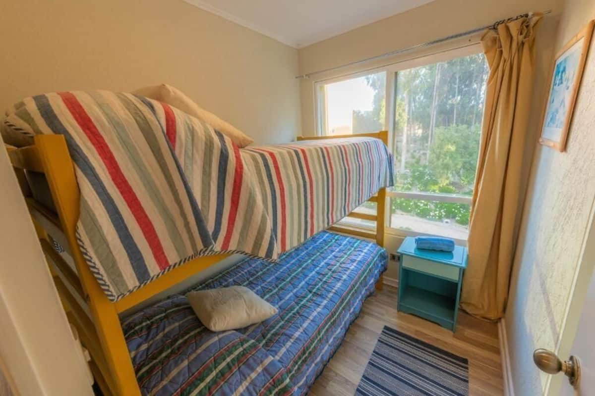 Une chambre avec un lit superposé, une petite table, une fenêtre avec des rideaux et une vue sur la verdure.