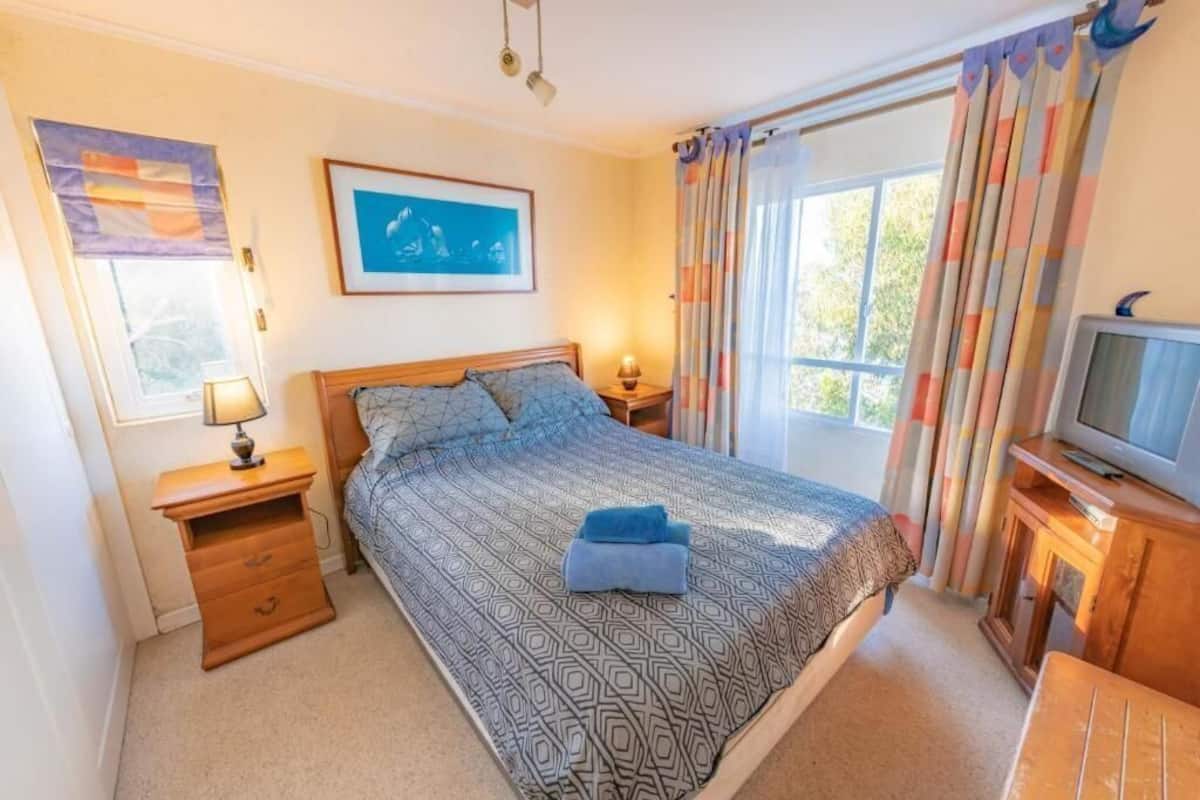 Une chambre à coucher avec un lit, une table de chevet avec une lampe, une télévision et une fenêtre avec des rideaux.