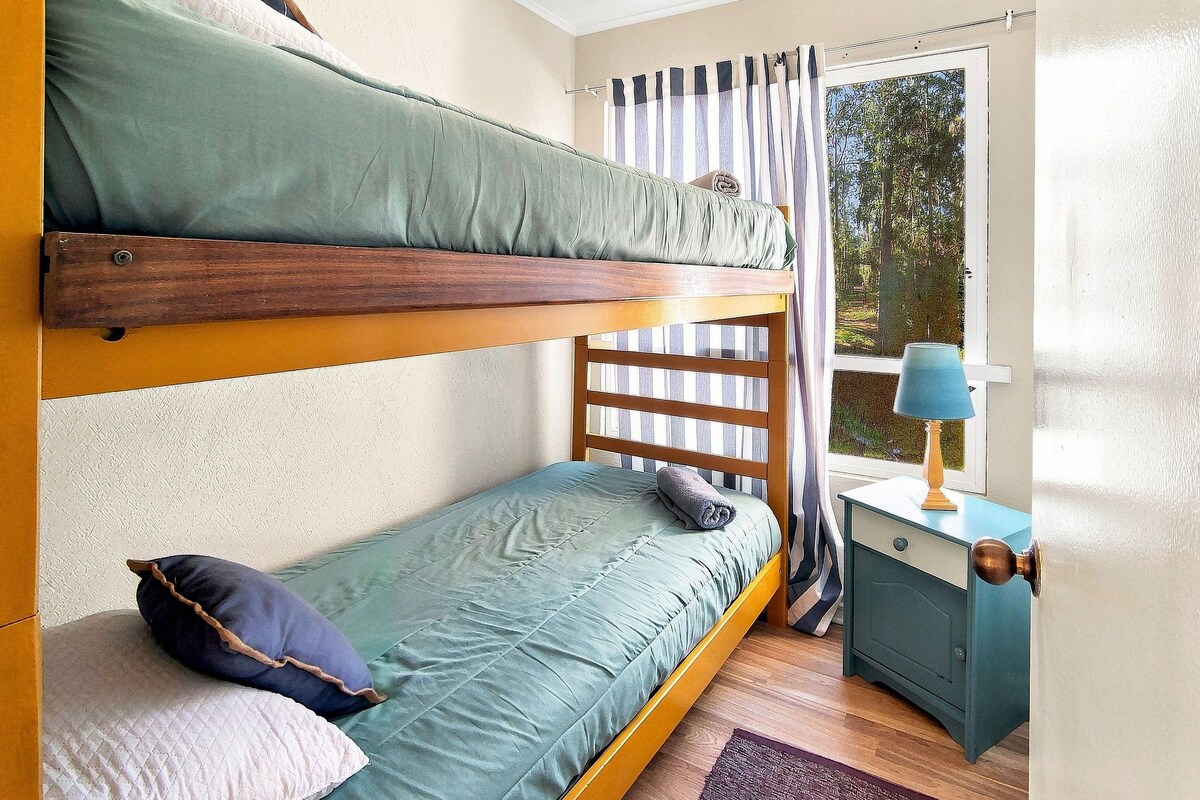 A bunk bed with a wooden frame, a single bed with a green mattress, a small nightstand with a lamp, and a window with curtains.