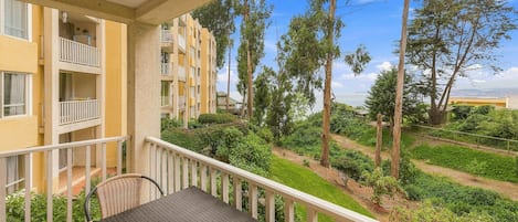 A balcony with a table and chairs, overlooking a landscaped area with trees and a distant view of a building.