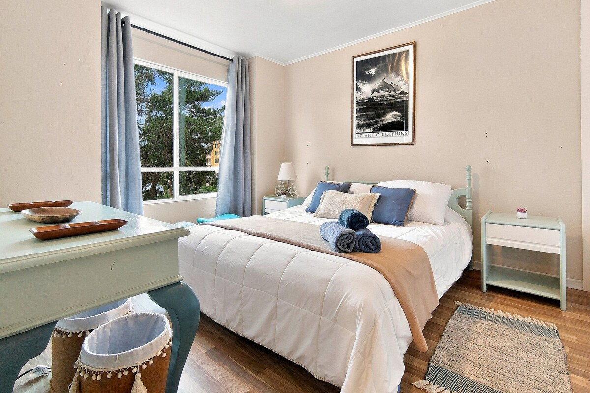 Un dormitorio con cama, mesitas de noche, una ventana con cortinas y un cuadro en la pared.