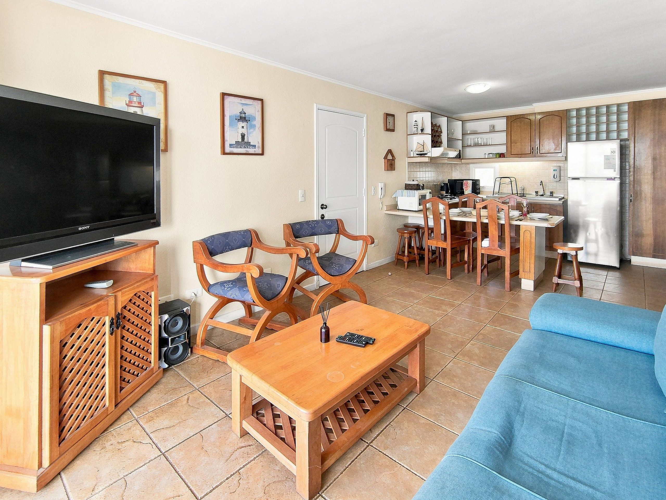A living room with a flat-screen TV, wooden furniture, a blue sofa, and a kitchen area with cabinets and appliances.
