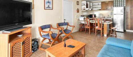 A living room with a flat-screen TV, wooden furniture, a blue sofa, and a kitchen area with cabinets and appliances.