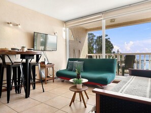 Una habitación con televisión montada en la pared, un sofá, una mesa de comedor con sillas y vista al mar.