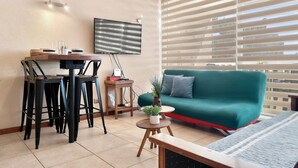 Una sala de estar con un sofá verde, una mesa de comedor con sillas, un televisor montado en la pared y una ventana con persianas.
