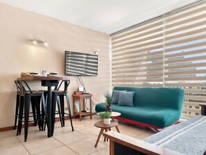 A living room with a green sofa, a dining table with chairs, a TV mounted on the wall, and a window with blinds.