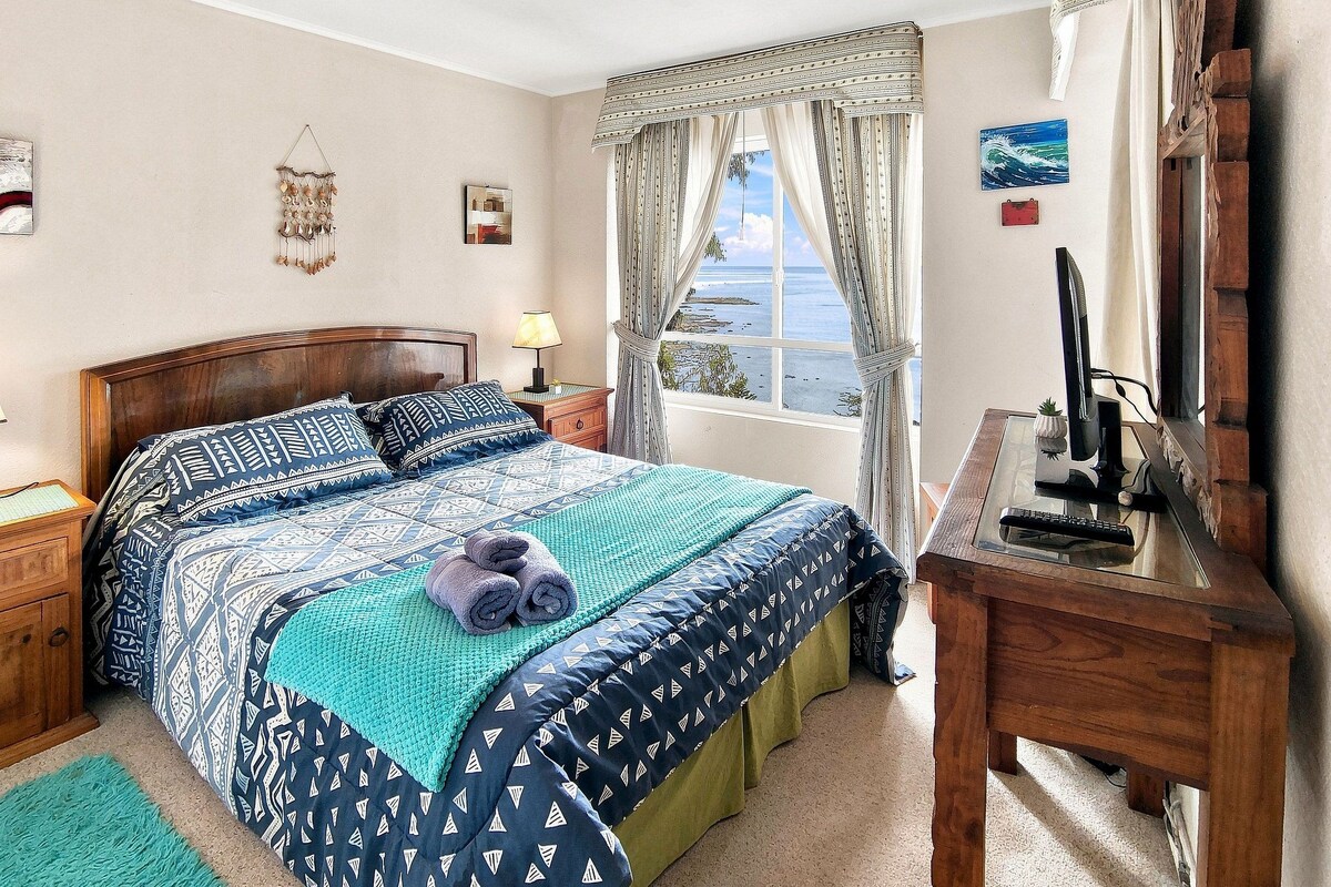 Une chambre à coucher comprenant un lit, une table de chevet, un bureau, une fenêtre avec des rideaux et une vue sur la mer.