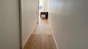 Un couloir étroit, doté d’un plancher en bois et de murs blancs, mène à une pièce où se trouvent un canapé de couleur sombre et une petite table.