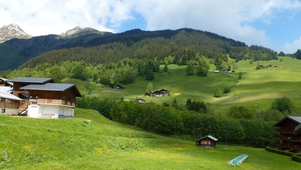 Un paysage rural avec des maisons sur une colline, une piscine et des montagnes en arrière-plan.