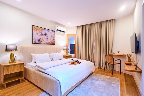 A modern hotel room with a large bed, a bedside table with a lamp, a small table with a wine glass and a vase, a chair, and a TV mounted on the wall.