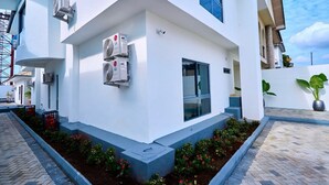 A modern building with air conditioning units mounted on the exterior wall, a glass door entrance, and a paved pathway with flower beds.