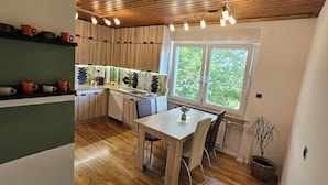 A kitchen with wooden cabinets, a dining table, chairs, a window with a view of greenery, and a shelf with cups.
