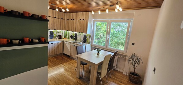 A kitchen with wooden cabinets, a dining table, chairs, a window with a view of greenery, and a shelf with cups.