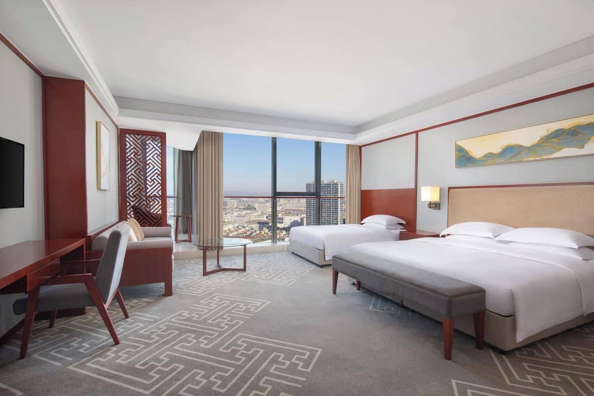 A hotel room with a large bed, a desk, a chair, and a view of the cityscape through large windows.