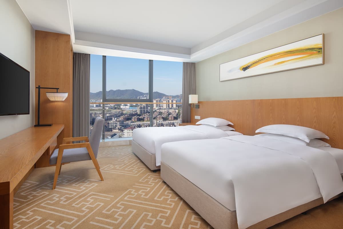 A hotel room with two beds, a desk, a chair, and a large window offering a city view.