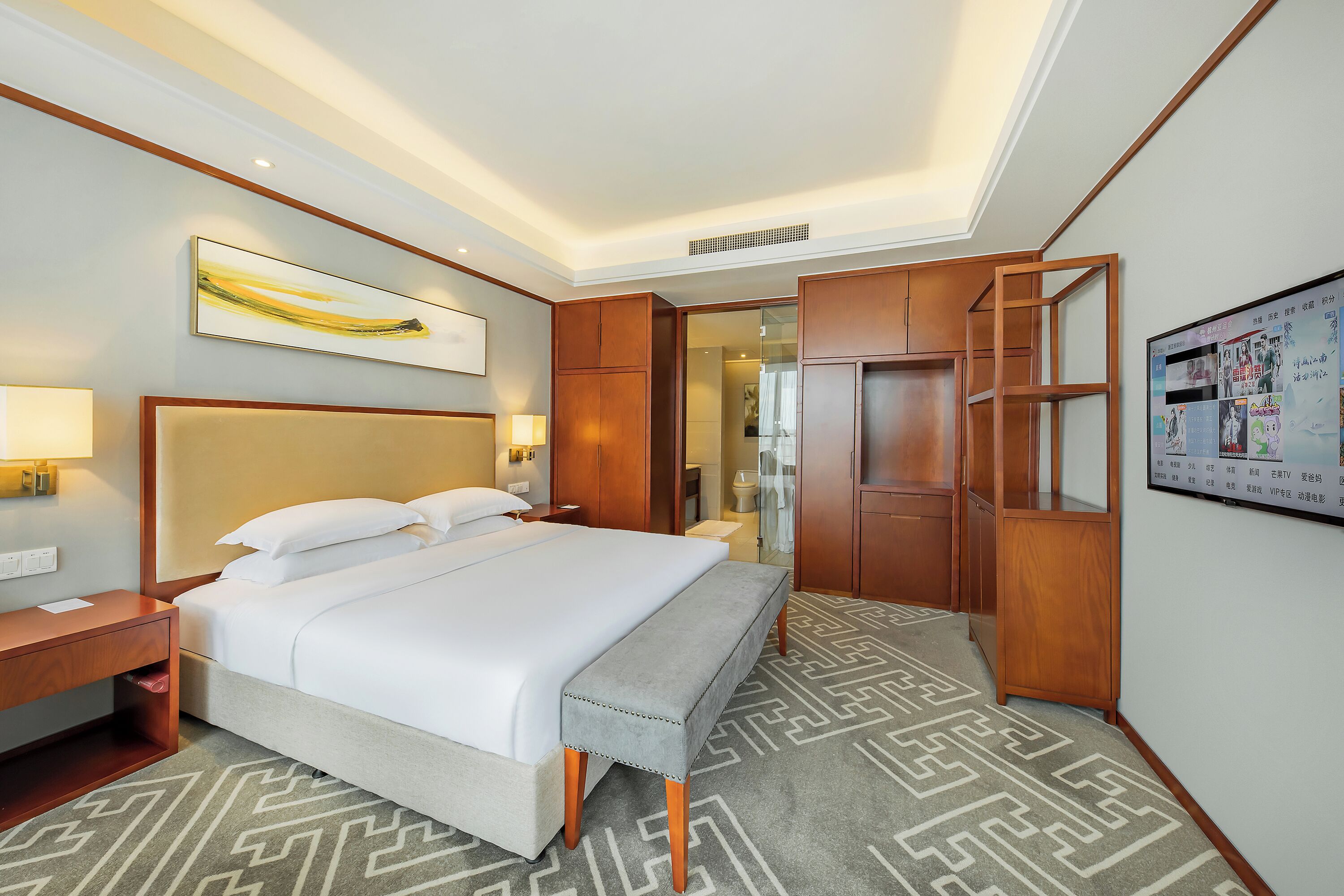Habitación de hotel con una cama grande, mesitas de noche, un banco, armario y televisor montado en la pared.