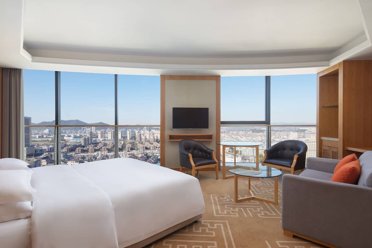 A hotel room with a large bed, two armchairs, a sofa, a TV, and a city view.