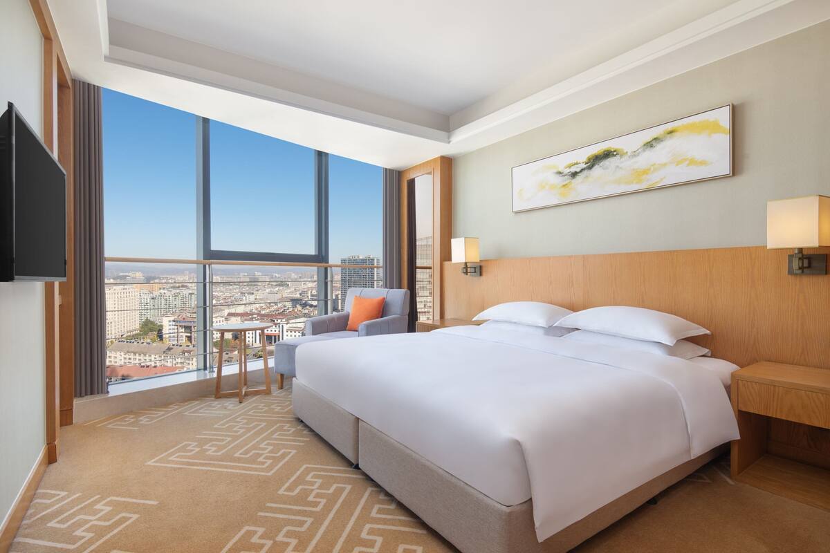 A hotel room with a large bed, a TV, a desk, a chair, and a view of the city.