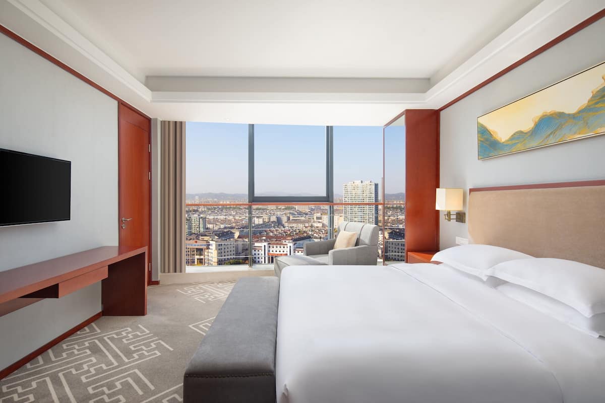 A hotel room with a large bed, a TV, a desk, and a view of a cityscape through large windows.