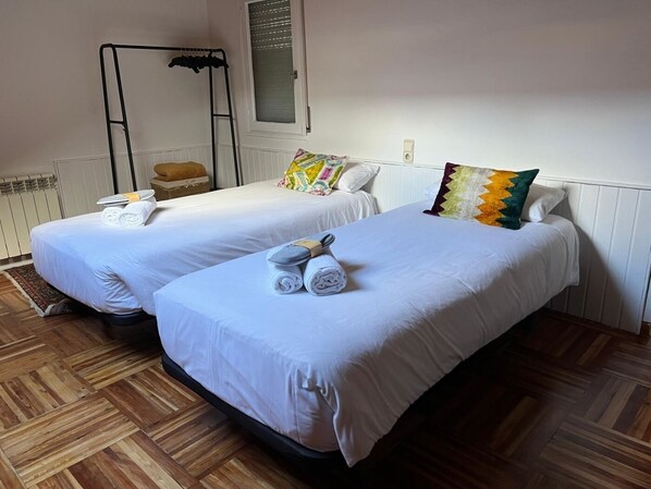 Une chambre d’hôtel avec deux lits simples, chacun équipé de draps et de serviettes blancs, un parquet en bois, une fenêtre avec des stores et une petite table avec une chaise.