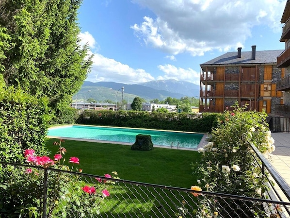 A pool surrounded by greenery, a building with balconies, and mountains in the background.