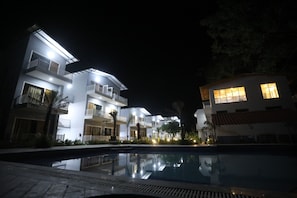 A modern hotel with a swimming pool, multiple balconies, and well-lit exterior.