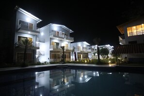 A modern hotel with multiple balconies, a swimming pool, and well-lit outdoor areas.