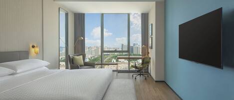 Ein modernes Hotelzimmer mit einem großen Bett, einem Schreibtisch mit Stuhl, einem an der Wand befestigten Fernseher und Blick auf die Stadt durch große Fenster.
