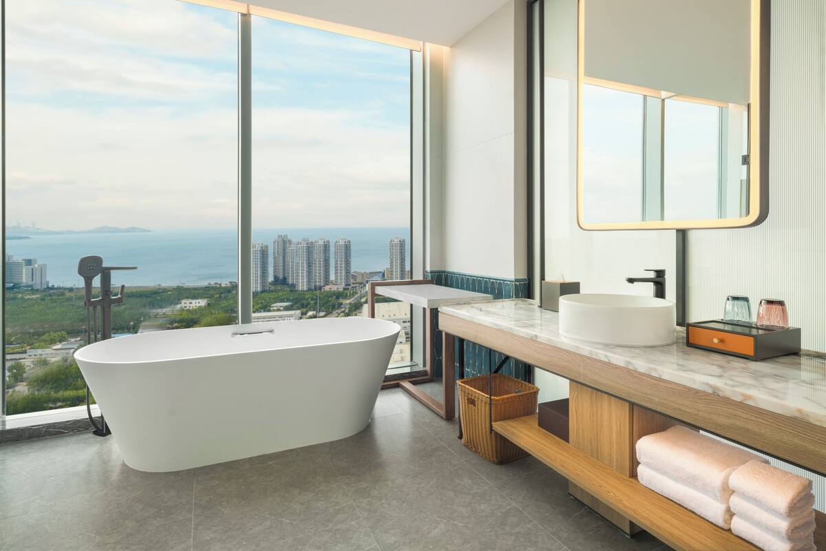 A modern bathroom with a freestanding bathtub, a large mirror, and a view of a cityscape through large windows.