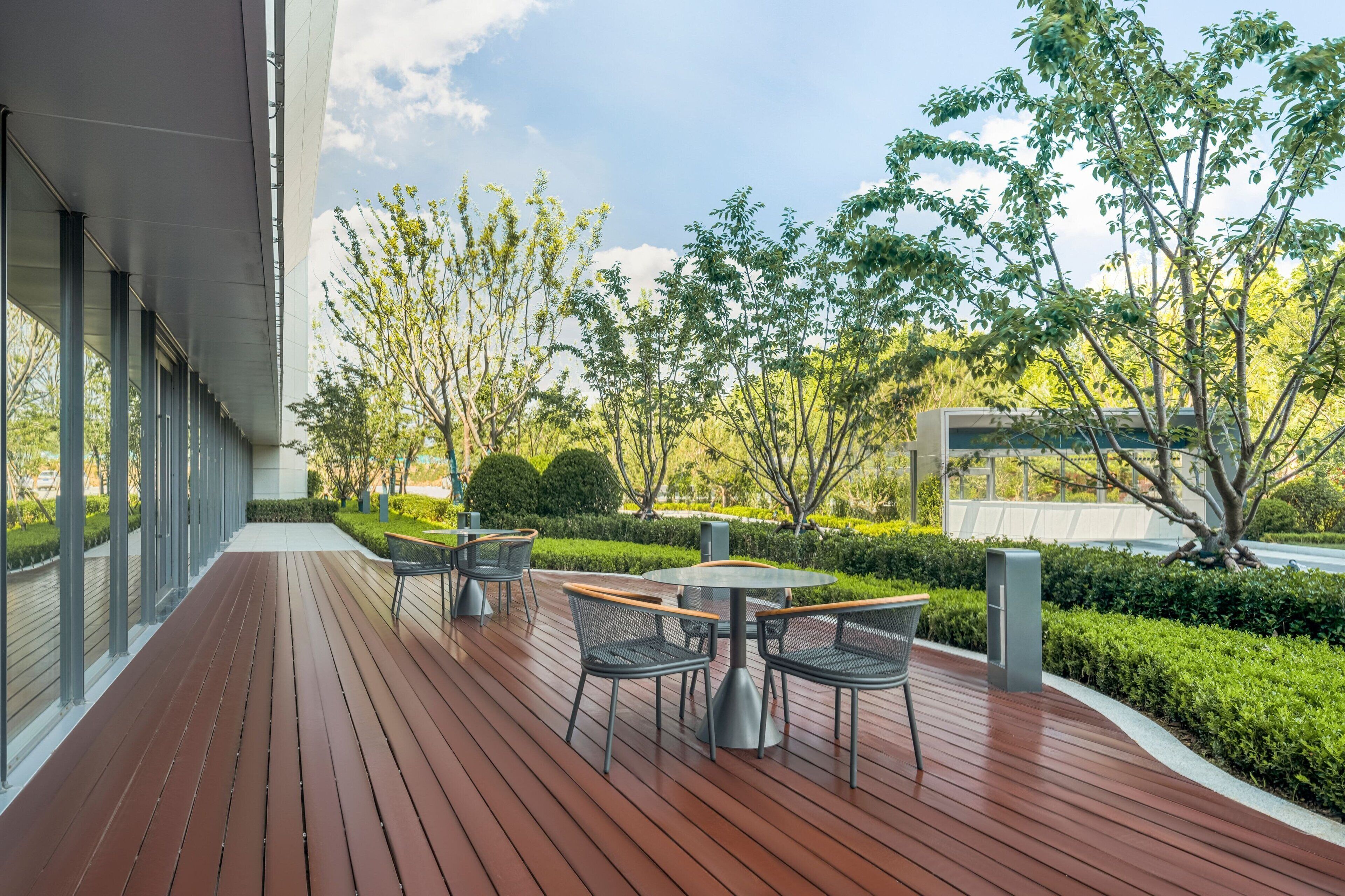 An outdoor patio with wooden flooring, metal chairs, and tables, surrounded by greenery and trees.
