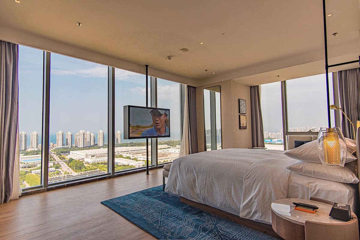 A modern hotel room with a large bed, a TV mounted on the wall, and a city view through large windows.