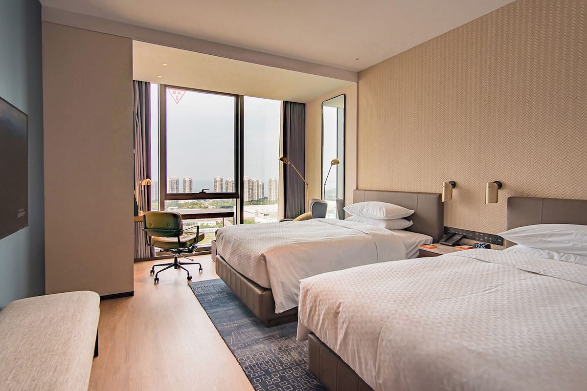 A hotel room with two beds, a desk, a chair, a large window with a city view, and a mirror.
