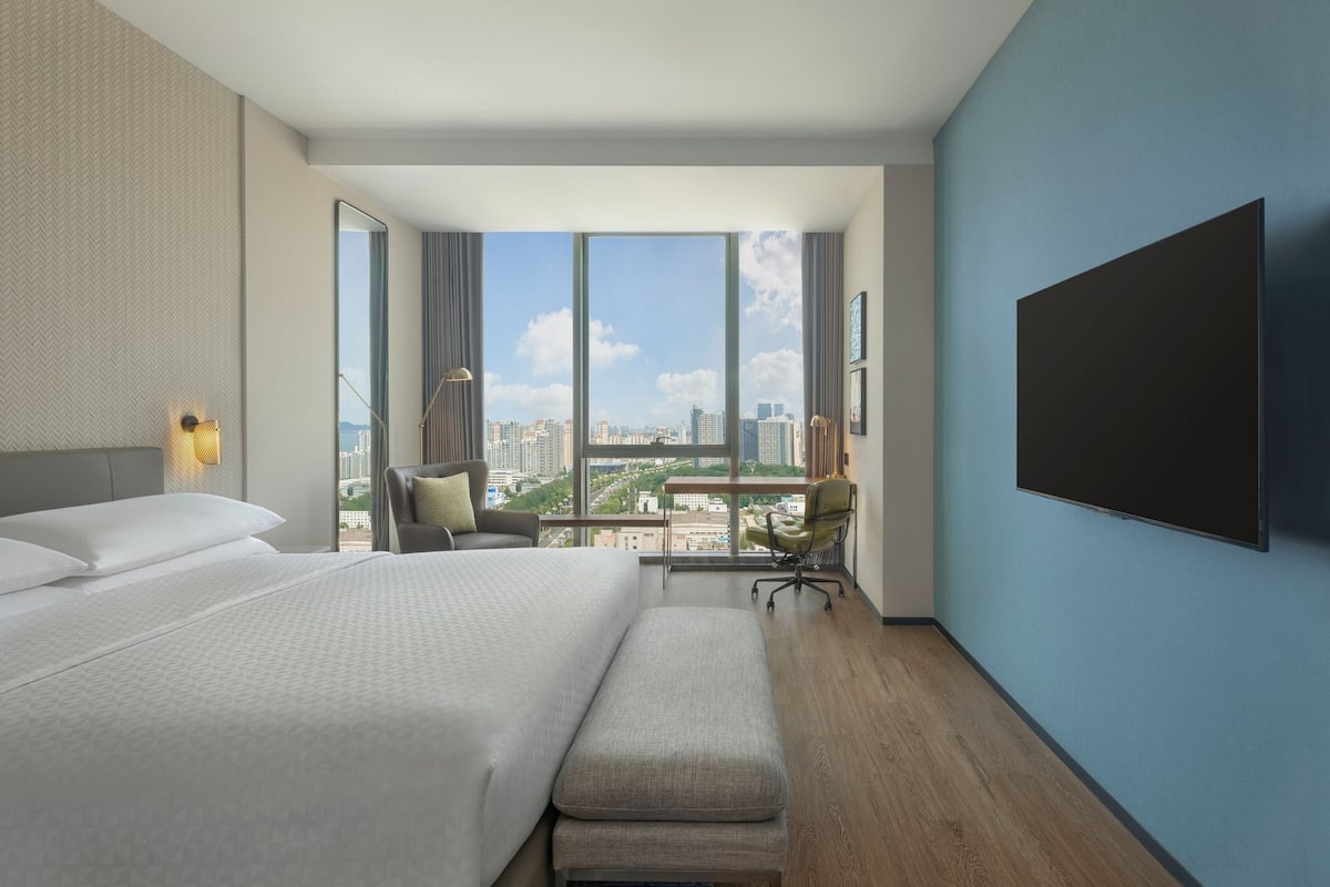 A modern hotel room with a large bed, a desk, and a view of the cityscape through large windows.