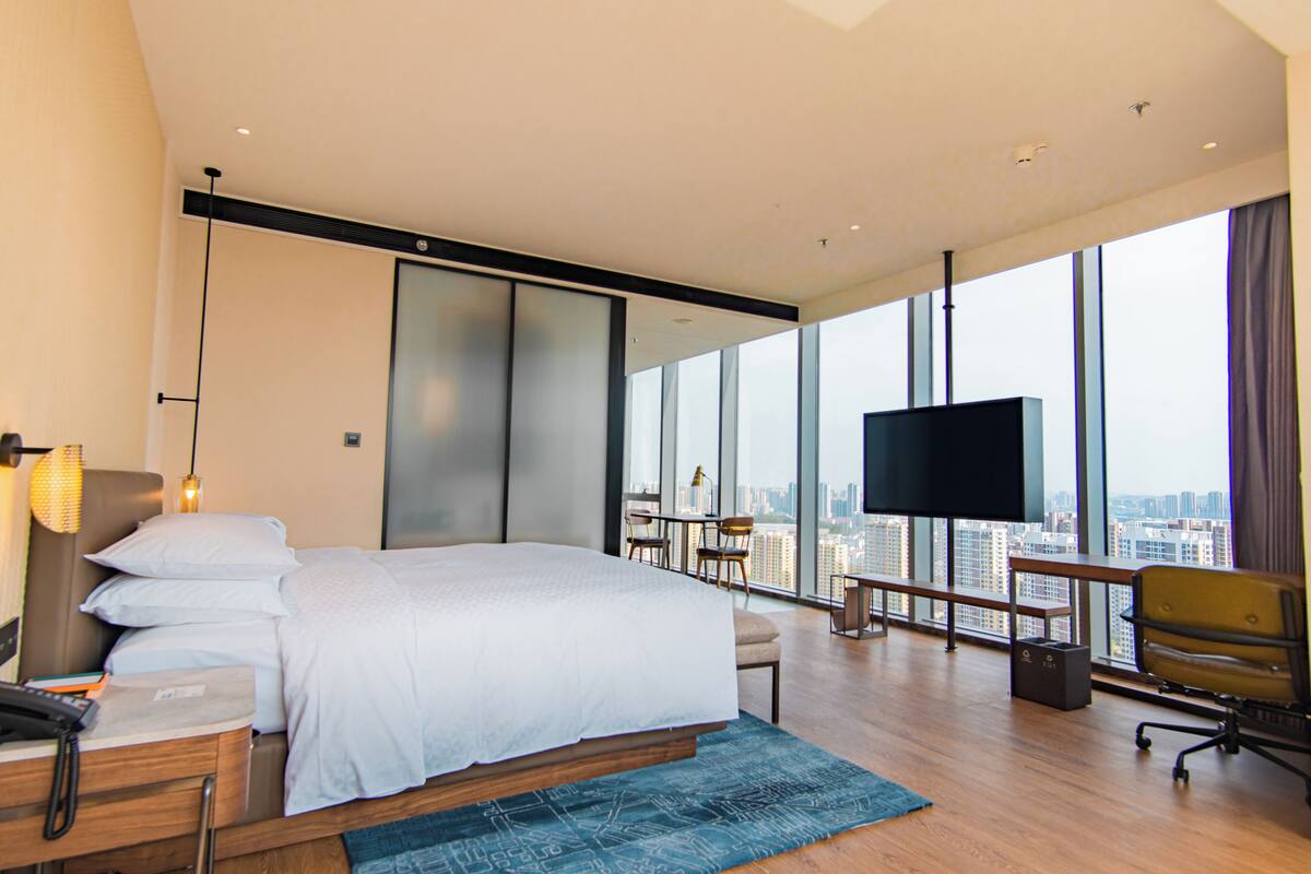 A modern hotel room with a large bed, a desk with a phone, a TV, and a city view through large windows.