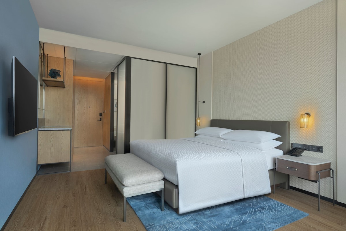A modern hotel room with a large bed, a bedside table with a lamp, a flat-screen TV, and a small seating area.