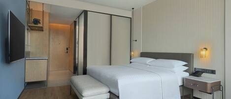 A modern hotel room with a large bed, a bedside table with a lamp, a flat-screen TV, and a small seating area.