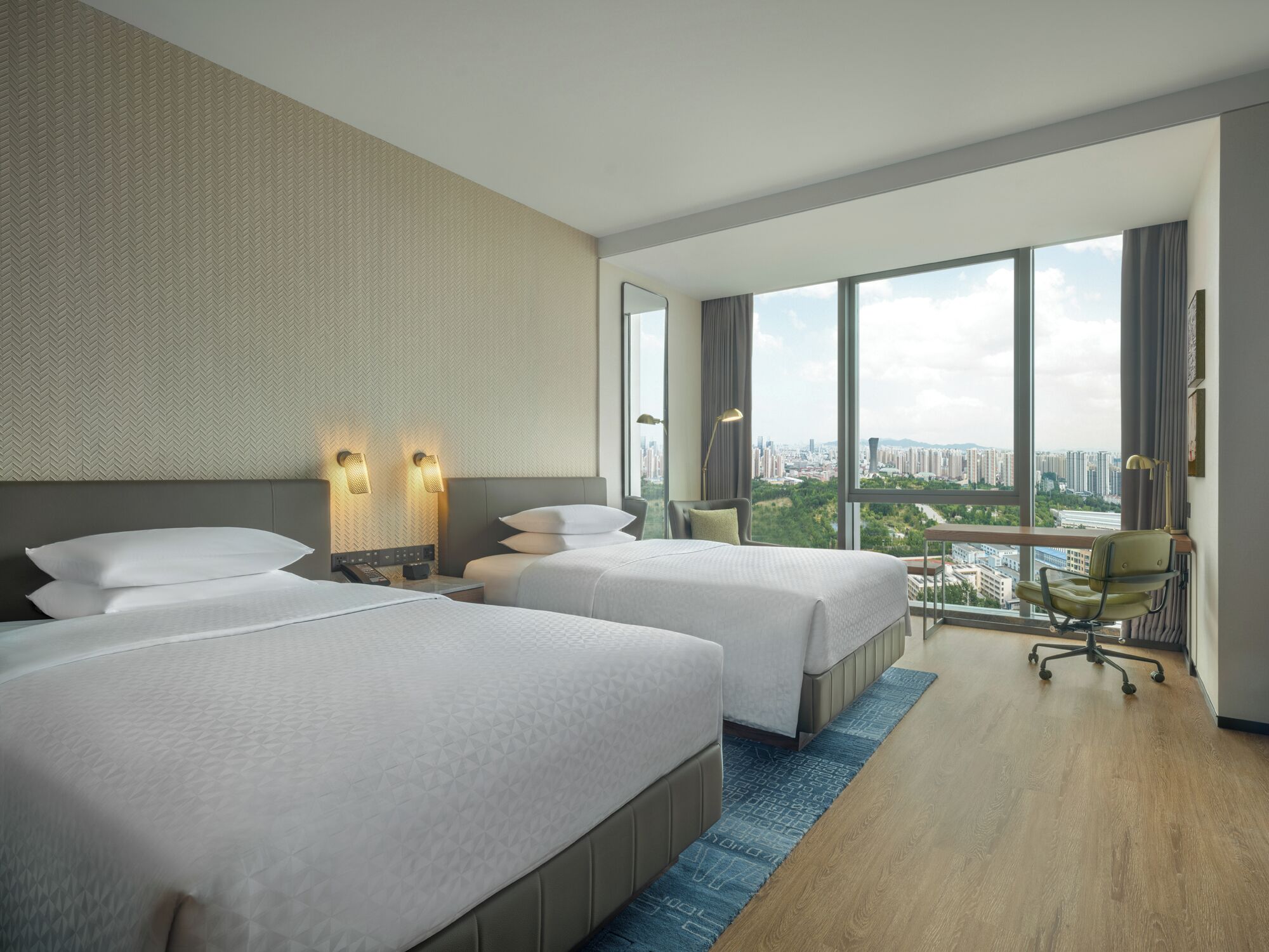 A hotel room with two beds, a large window with a city view, and a desk with a chair.