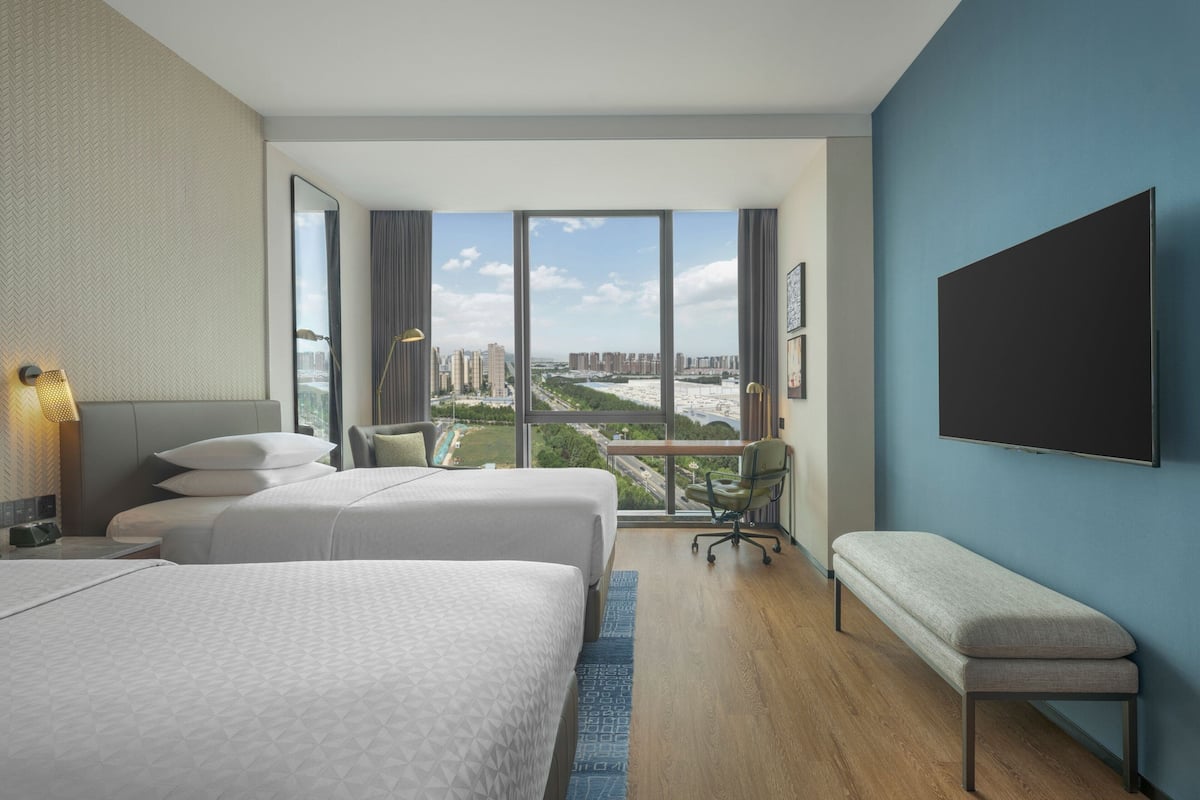 A modern hotel room with two beds, a large window offering a city view, a bench, and a flat-screen TV mounted on the wall.