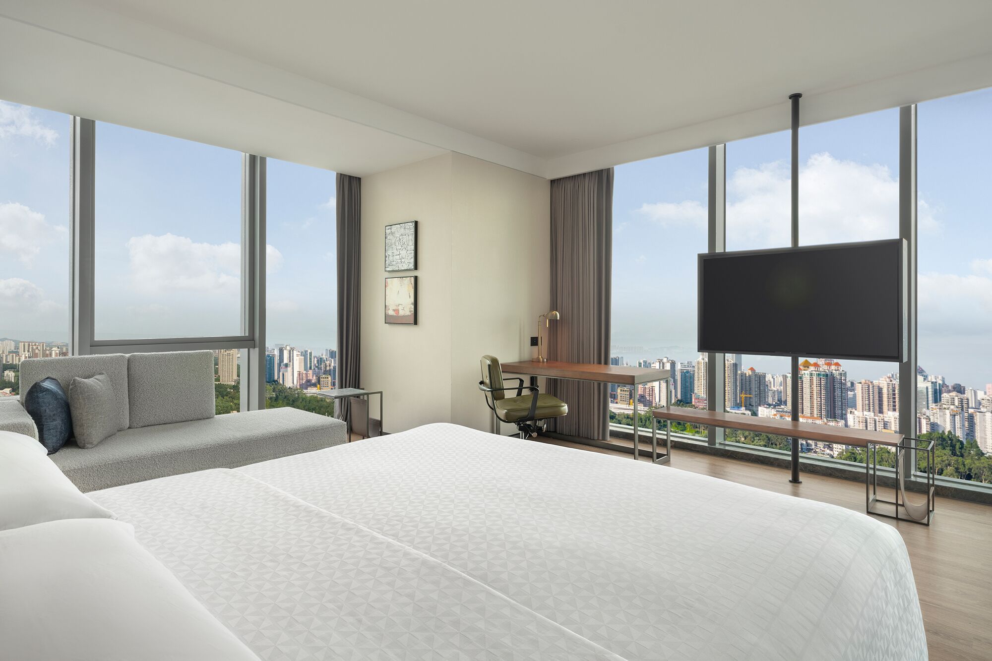 A modern hotel room with a large bed, a desk, a chair, and a TV. The room has large windows offering a city view.